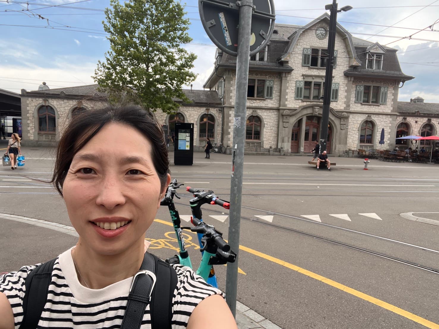 Dr. Mari smiling outside the Zürich station, with trams and an old stone building behind her